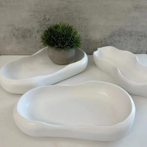 May include: Three white, organically shaped ceramic trays. One tray holds a small, round, gray pot with a green plant. The trays have smooth, curved edges and a glossy finish. The trays are empty and ready to be used for decorative purposes.