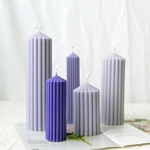May include: A collection of five pillar candles in varying shades of purple and lavender. The candles have a ribbed design and are arranged on a white surface. The candles are of different heights and shapes, creating a visually appealing display.