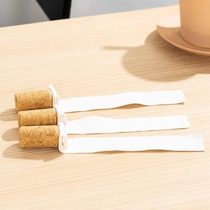 May include: Three cylindrical cork stoppers arranged in a vertical stack, each featuring a long, flat, white fabric ribbon attached to one end. The corks have a natural tan, porous texture, while the attached white ribbons extend horizontally.