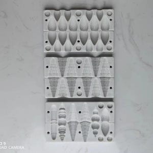 以下が含まれることがあります： 涙型、円錐形、装飾オーナメントなど、さまざまな形状の3つのグレーのシリコン型。各型には、一度に複数のアイテムを作成するための複数のキャビティがあります。型は白い大理石の表面に配置されています。