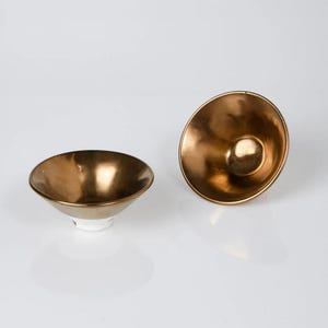 May include: Two small, gold-colored bowls with a metallic sheen. One bowl is upright, while the other is tilted on its side. The bowls have a white base and a rounded shape, suitable for serving small portions.