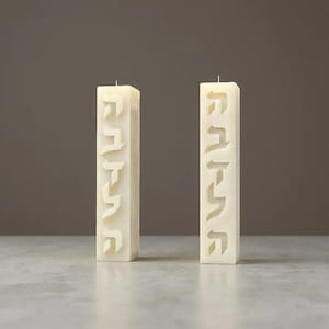 Può includere: Due candele rettangolari color avorio con lettere ebraiche intagliate. Le candele sono in posizione verticale su uno sfondo neutro. Le candele hanno un design semplice ed elegante, adatto per l'arredamento della casa o occasioni speciali.
