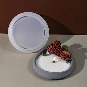 May include: A round, gray concrete bowl containing a white candle with a lit wick, decorated with several dried red roses. Beside the candle sits a matching circular, translucent silicone mold used for creating the bowl's lid or base.