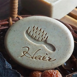 May include: Oval-shaped, pale green soap with a comb design and the word "Hair" etched into the surface. The soap is speckled and sits in a woven basket. Another bar of soap is visible in the background.