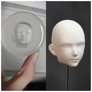 May include: A clear, circular mold with a detailed face cast inside, next to a white doll head on a metal rod. The doll head has a smooth surface with defined features, including eyes, nose, and ears. The mold is held by a hand.