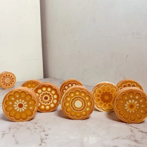 Pu&ograve; includere: Una collezione di pezzi decorativi rotondi in ceramica con un design floreale. I pezzi presentano una calda tavolozza di colori arancione, giallo e bianco, con motivi intricati. Gli articoli sono disposti su una superficie di marmo.