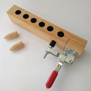 Pode incluir: Um gabarito de madeira clara com seis orifícios forrados a preto e duas rolhas de cortiça. Uma braçadeira de metal com uma pega vermelha está presa ao bloco de madeira. Projetado para projetos de carpintaria ou artesanato.
