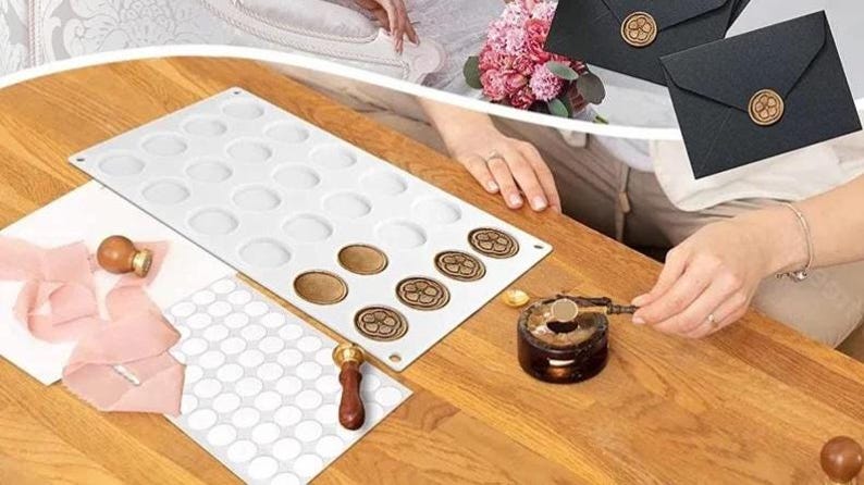 Op de afbeelding: Een witte siliconen mal met 28 ronde holtes voor het maken van waszegels. Een houten handvat met een metalen punt wordt gebruikt om was te smelten en op de zegels aan te brengen. Een vel wit papier met 50 ronde stickers is ook inbegrepen.