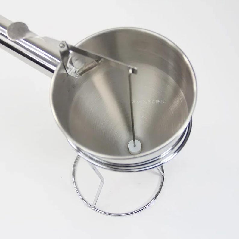 Puede incluir: Un dispensador de masa de acero inoxidable con un embudo, un mango y un soporte. El dispensador tiene una varilla de metal que atraviesa el embudo.