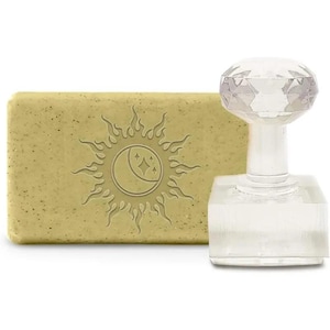 Peut inclure: Un pain de savon rectangulaire vert clair avec un motif de soleil et de lune, à côté d'un tampon transparent et facetté. Le tampon a une base carrée et un dessus en forme de diamant. Le savon a une texture tachetée.