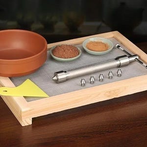 May include: A wooden tray containing a terracotta bowl, a yellow plastic scraper, two small ceramic dishes with brown powder, and a silver metal extruder tool with five interchangeable nozzles arranged in a row on the tray's gray mesh surface.