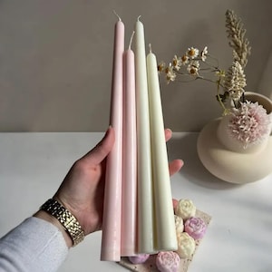 Puede incluir: Una mano sostiene cuatro velas cónicas en tonos rosa y crema. Las velas son largas y delgadas, con un acabado liso y mate. El fondo incluye un jarrón con flores y detalles florales decorativos.