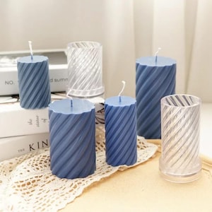 Può includere: Diverse candele blu a spirale con stoppini bianchi sono esposte accanto a stampi per candele cilindrici trasparenti. Le candele sono di varie dimensioni e poggiano su un centrino di pizzo bianco e libri. Le candele hanno un design strutturato e scanalato.