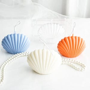May include: Three shell-shaped candles in light blue, white, and orange, each featuring a central wick. A clear, empty shell-shaped candle mold sits behind them, and a string of white faux pearls is draped across the foreground.