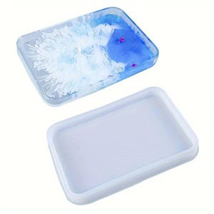 Puede incluir: Una bandeja rectangular de resina con un patrón de remolinos de pigmentos blancos y azules con pequeños acentos rosas incrustados, junto con un molde de silicona blanco translúcido a juego para fundir la forma rectangular.