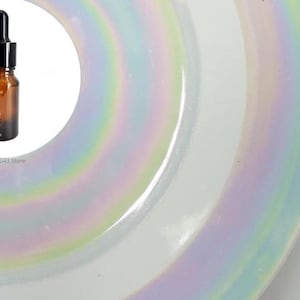 以下が含まれることがあります： 黒いドロッパー付きの透明なガラス瓶で、中には茶色の液体が入っています。瓶の側面には目盛りがあり、ミリリットルで表示されています。瓶は虹色の虹色の模様が施された白い表面の上に置かれています。