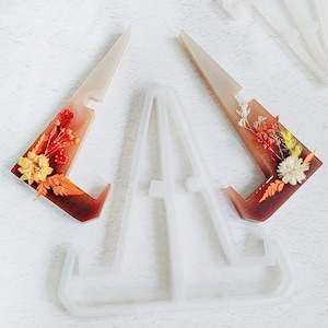 May include: A clear silicone mould and two resin phone stands. The resin stands are decorated with dried flowers in shades of yellow, orange, and brown. The stands have a light brown and clear colour scheme.