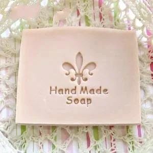 Puede incluir: Una pastilla de jabón rectangular de color beige claro con un símbolo de flor de lis en relieve centrado sobre el texto Hand Made Soap. El jabón tiene una superficie lisa y mate con bordes rectos y limpios de diseño sencillo.