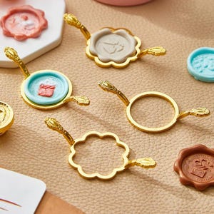May include: An assortment of gold-coloured wax seal stamp handles with ornate detailing. Several wax seals in shades of pink, blue, and brown are also present. The seals display detailed embossed patterns, indicating a stationery or craft theme.