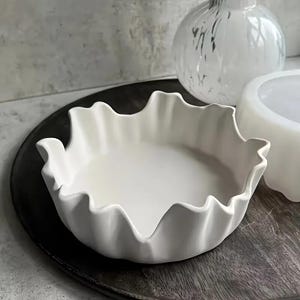 May include: A white ceramic bowl with a ruffled edge sits on a dark wooden tray. The bowl has a shallow, round shape. A white vase and a white decorative bowl are in the background.