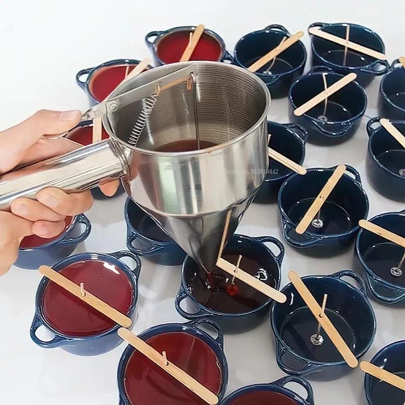 Puede incluir: Una persona vierte cera roja en peque&ntilde;os recipientes de cer&aacute;mica azul con mechas de madera. La cera se vierte desde un embudo de acero inoxidable con un mango de resorte.