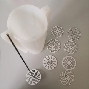 Pu&ograve; includere: Uno stampo bianco di forma astratta con un'asta di metallo e un design geometrico circolare all'estremit&agrave;. Diversi sottobicchieri bianchi tagliati al laser con vari motivi geometrici sono sparsi intorno allo stampo. I sottobicchieri presentano disegni floreali, a stella e a spirale.