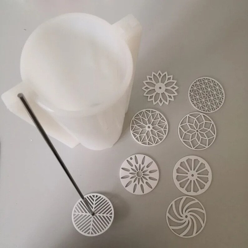 Pu&ograve; includere: Uno stampo bianco di forma astratta con un'asta di metallo e un design geometrico circolare all'estremit&agrave;. Diversi sottobicchieri bianchi tagliati al laser con vari motivi geometrici sono sparsi intorno allo stampo. I sottobicchieri presentano disegni floreali, a stella e a spirale.