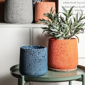 May include: A set of cylindrical plant pots with a porous, textured surface. One pot is dark blue, one is orange, and one is light gray. The orange pot contains a green leafy plant and sits on a circular base. The pots have a rough, stone-like finish.