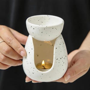 May include: A white ceramic essential oil diffuser with black speckles. The diffuser has a top bowl and a lower chamber with a lit candle inside. The design is a modern, minimalist style, perfect for aromatherapy.