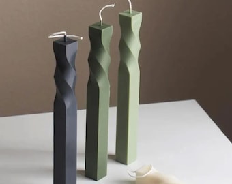 May include: A collection of decorative candles in various colors, including dark gray and shades of green. The candles have a twisted, geometric design and are arranged on a white surface. The wicks are visible at the top of each candle.