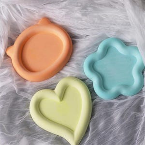 May include: Three decorative trays in different shapes and colors. An orange circular tray with two small protrusions, a light blue cloud-shaped tray, and a yellow heart-shaped tray. All trays have a matte finish and are set against a white fabric background.