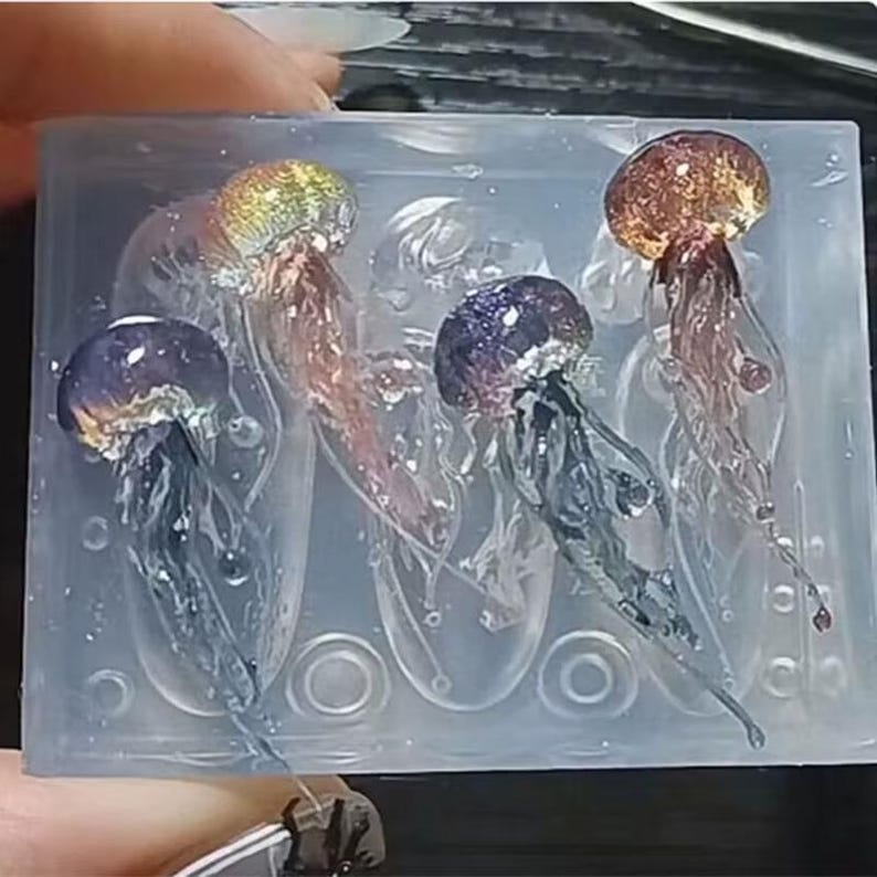 Puede incluir: Un molde de silicona transparente con cuatro dise&ntilde;os de medusas. Las medusas tienen gorras iridiscentes coloridas y cuerpos transl&uacute;cidos en tonos de rosa, morado y azul. El molde est&aacute; sostenido por una mano.