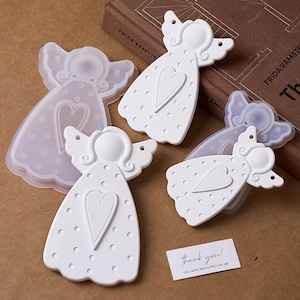 Puede incluir: Moldes y productos terminados blancos en forma de ángel con detalles de corazón y puntos. La imagen incluye una tarjeta de agradecimiento y un libro. Los moldes son de un material translúcido, mientras que los ángeles terminados son blancos.
