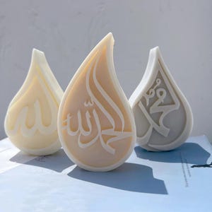 May include: Three teardrop-shaped decorative objects in cream, beige, and light gray. Each piece features raised white Arabic calligraphy embossed on the front surface. The objects have a smooth, solid finish and a distinct pointed top design.