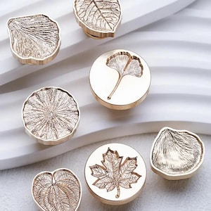 Może przedstawiać: Kolekcja złotych stempli do wosku z różnymi wzorami liści. Stemple przedstawiają szczegółowe odciski liści, w tym liścia miłorzębu, liścia klonu i innych wzorów botanicznych. Stemple są ułożone na białej powierzchni.