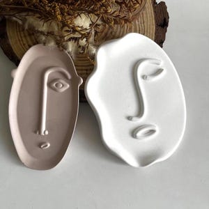 Puede incluir: Dos bandejas decorativas con forma de rostro. Una es de color rosa claro, la otra blanca. Ambas bandejas tienen un diseño minimalista con rasgos faciales en relieve, incluyendo ojos, narices y bocas. Las bandejas probablemente son de cerámica o material similar.