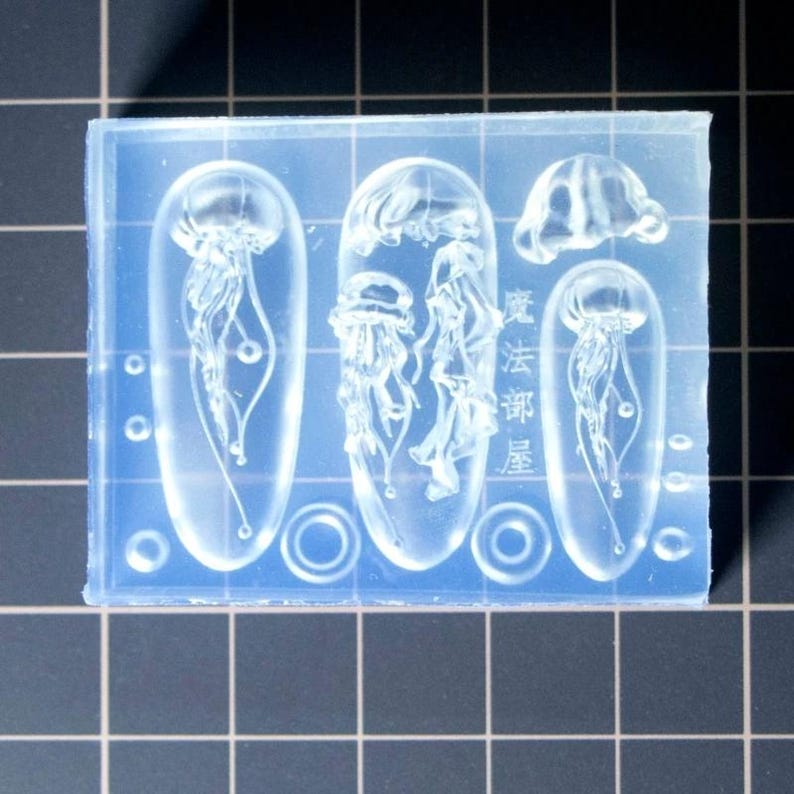 Puede incluir: Un molde de silicona transparente y rectangular con dise&ntilde;os de medusas. El molde incluye tres formas ovaladas alargadas con medusas, una peque&ntilde;a medusa y el texto japon&eacute;s "Magic Department Room".