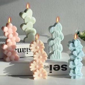 Puede incluir: Cinco velas decorativas en varios colores pastel, incluyendo rosa, verde y azul. Cada vela tiene un diseño único, como corazones apilados, flores y patrones ondulados. Las velas están encendidas, con llamas visibles en la parte superior.