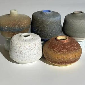 May include: Five small ceramic vases in various colors and textures. The vases are all round and have a small opening at the top. They are arranged in a row on a white surface.