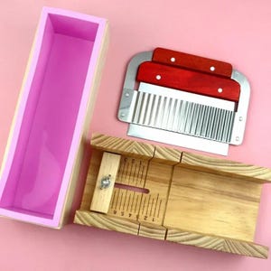 May include: A soap-making kit with a pink silicone mold inside a wooden box, a wooden soap cutter with measurements, and a stainless steel wavy soap cutter with red wooden handles. The items are arranged on a pink surface.