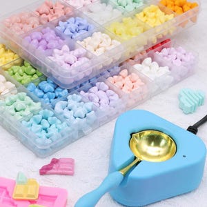 May include: A clear plastic storage container with many compartments filled with small, star-shaped wax beads in various pastel colors. A light blue electric wax melting furnace with a gold-colored metal spoon and a pink silicone mold are nearby.