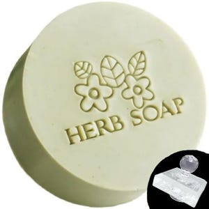Puede incluir: Jabón de hierbas redondo, verde claro, con diseño floral y la inscripción "HERB SOAP". Un sello transparente con una parte superior de cristal es visible en la parte inferior derecha. El jabón tiene un aspecto natural y orgánico.