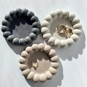 May include: Three decorative ring dishes in gray, white, and beige. Each dish has a rounded, bubble-like edge. The white dish holds two gold hoop earrings, and the beige dish holds a gold ring. The dishes are arranged on a white surface.
