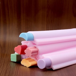 May include: A pink silicone mold with various colorful shapes, including a heart, flower, and square. The mold has a light blue and purple accent. The shapes are in shades of red, green, and orange. The background is a wooden surface.