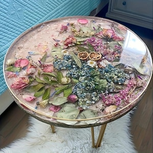 Può includere: Tavolino rotondo con piano in resina trasparente con fiori secchi nei toni del rosa, blu e viola. Il tavolo ha una struttura e gambe in metallo color oro. Il tavolo ha un diametro di circa 51 cm.