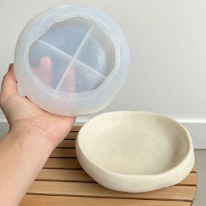 Op de afbeelding: Een heldere, ronde siliconen mal met vier secties, boven een kleine, crèmekleurige keramische kom gehouden. De kom heeft een gladde, afgeronde vorm. De items worden op een houten oppervlak gepresenteerd.