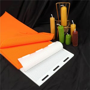 May include: A flexible orange silicone mold sheet with a hexagonal honeycomb pattern, partially overlapping a white rectangular silicone mold sheet with a matching honeycomb texture, alongside five cylindrical beeswax candles.