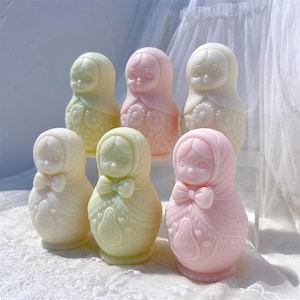 May include: A collection of six pastel-colored Matryoshka dolls. The dolls are in shades of pink, yellow, and white, with detailed floral and bow accents. Each doll has a distinct face and is arranged on a clear surface.