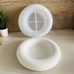 Puede incluir: Dos moldes circulares blancos para manualidades. Uno tiene forma de cuenco poco profundo, y el otro tiene un divisor en forma de cruz en el interior. Los moldes están hechos de un material liso y opaco, probablemente silicona o resina, y están diseñados para crear objetos decorativos.