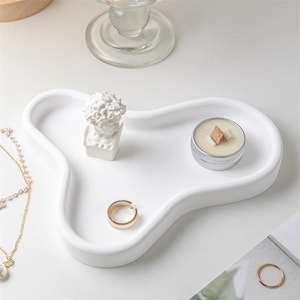 May include: A white, organically shaped tray holds a small bust, a gold ring, and a candle in a silver tin. A gold necklace and a glass candlestick are also visible. The items are arranged on a white surface.